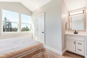 A bedroom with a bed, a window, a door, and a bathroom with a sink and mirror.