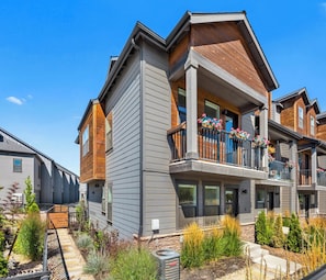 A multi-story residential building with a balcony, a small garden area, and a clear sky.