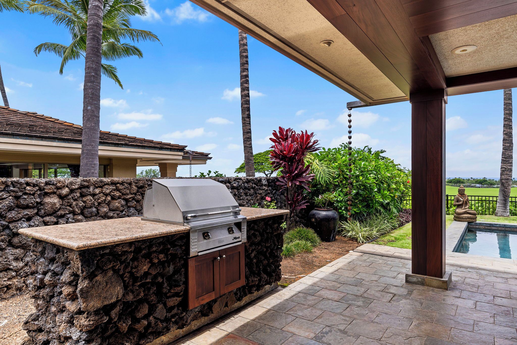 An outdoor kitchen area with a stone wall, a grill, and a pool in the background.