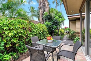 An outdoor patio with wicker furniture and a glass table, surrounded by lush greenery and palm trees. - Pakui Street Estate by Gather - Hualalai Golf Estate w/ Private Pool & Spa (Kailua Kona)