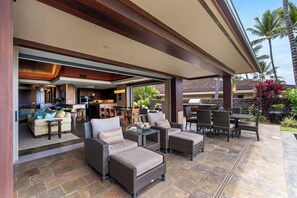 Ein Terrassenbereich im Freien mit Korbmöbeln, einem Esstisch und einer durchgehenden Tür erkennbaren Küche.