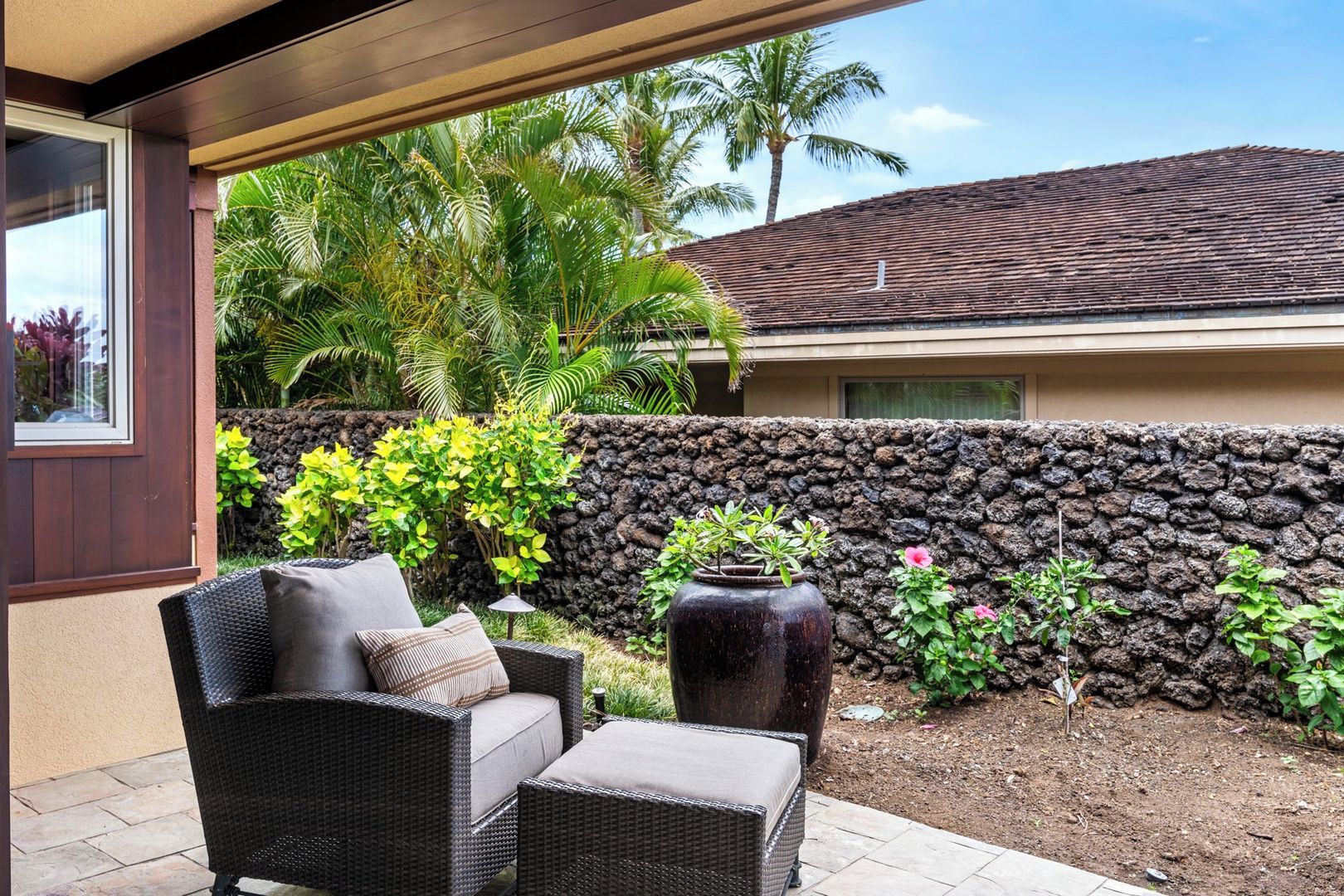 A patio with wicker furniture, a stone wall, and tropical plants.