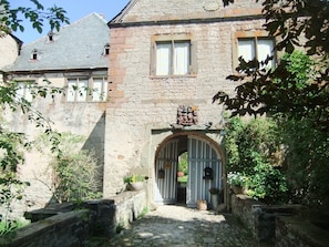 A stone building with a gated entrance, a coat of arms above the door, and a cobblestone path leading to the gate.