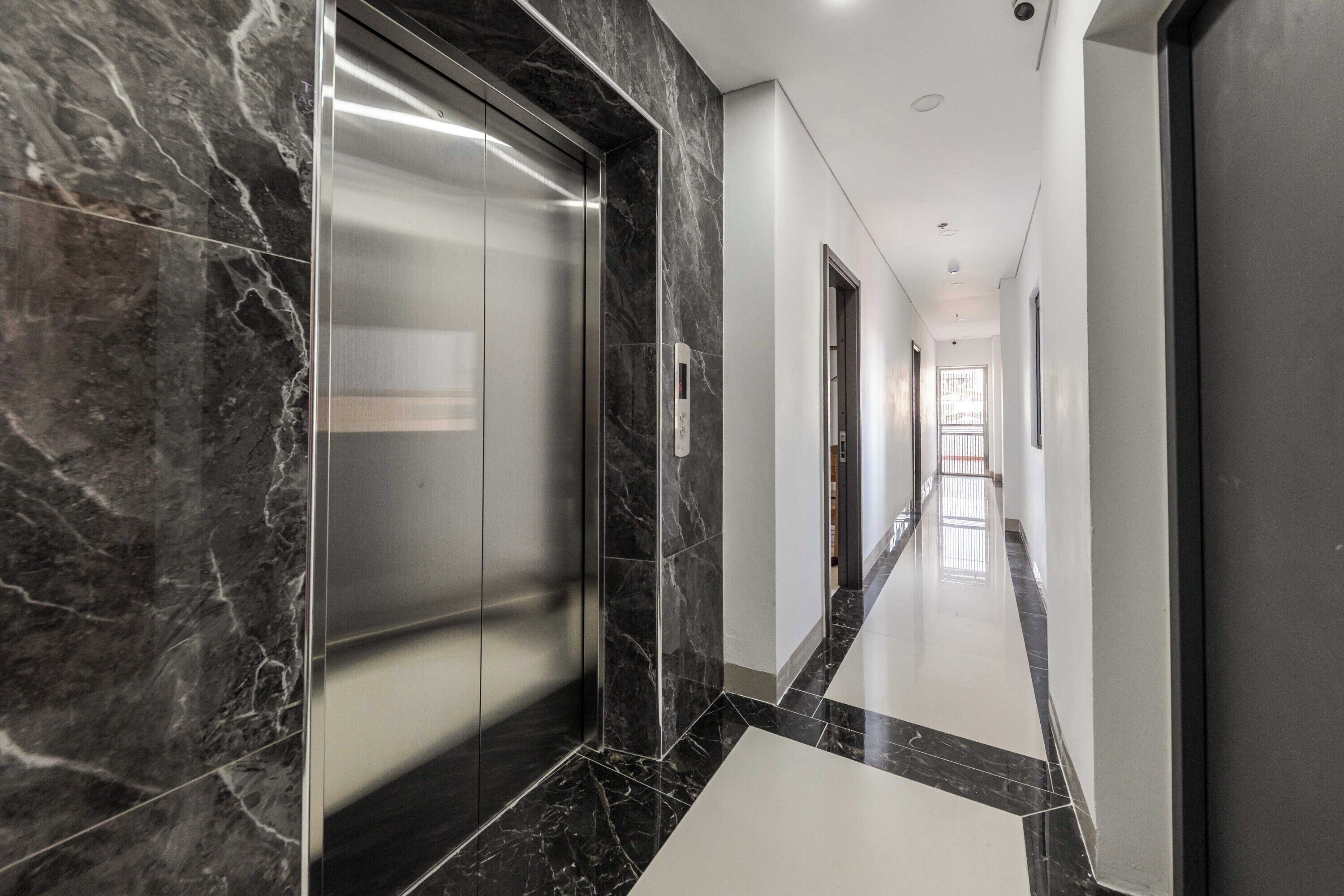 A modern hallway with marble flooring, a glass elevator, and a straight corridor leading to a bright room with a window.