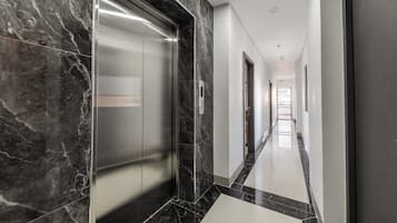 A modern hallway with marble flooring, a glass elevator, and a straight corridor leading to a bright room with a window.