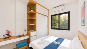 Un dormitorio con cama, escritorio, silla, estantería y ventana con vista a árboles.