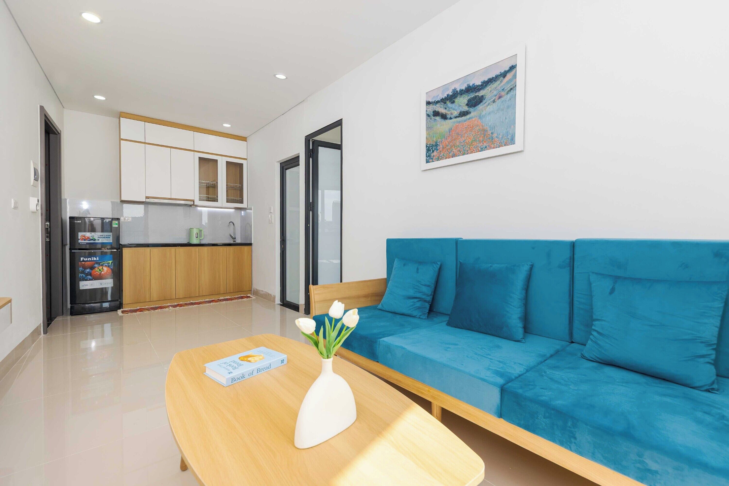 A modern living room with a blue sofa, a wooden coffee table with a book and a vase, and a kitchen in the background.