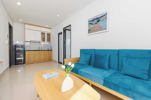 Una sala de estar moderna con un sofá azul, una mesa de centro de madera con un libro y un jarrón, y una cocina al fondo.
