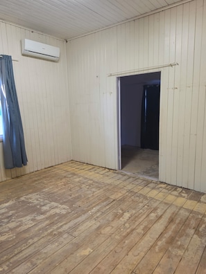 A room with wooden flooring, a white wall, and a door leading to another room.