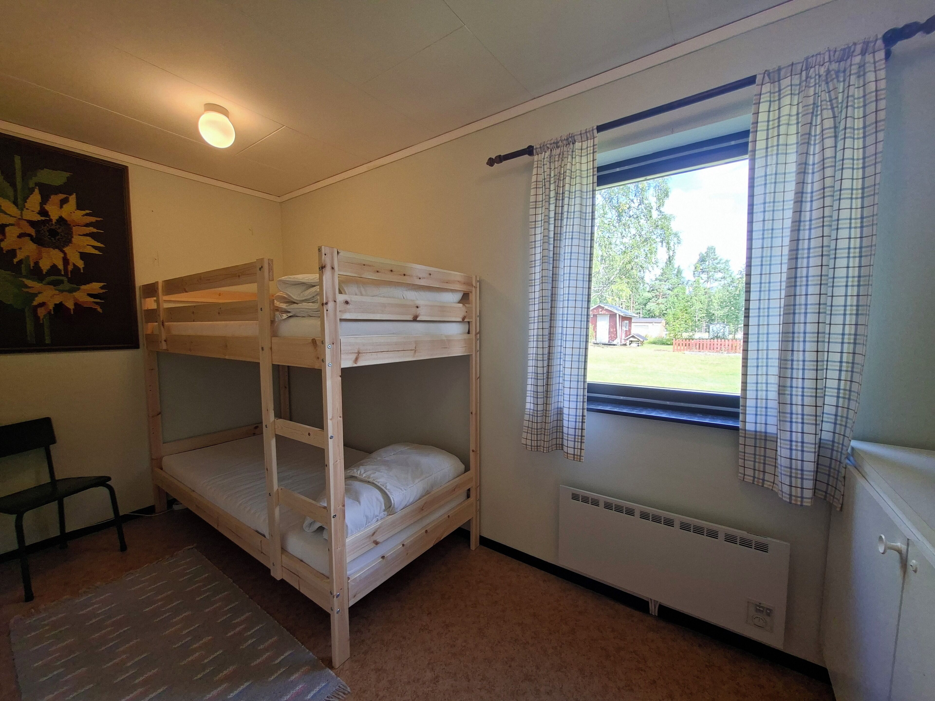 Une chambre avec un lit superposé, un radiateur, une fenêtre avec des rideaux, une chaise et un tableau représentant un tournesol.