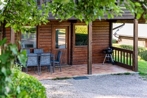 A wooden cabin with a patio area featuring a table and chairs, a grill, and a view of the surrounding greenery.