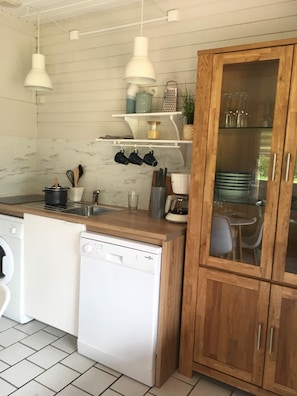 Une cuisine compacte équipée d’un lave-vaisselle, d’un meuble en bois aux portes vitrées, de tasses suspendues et d’une machine à café.
