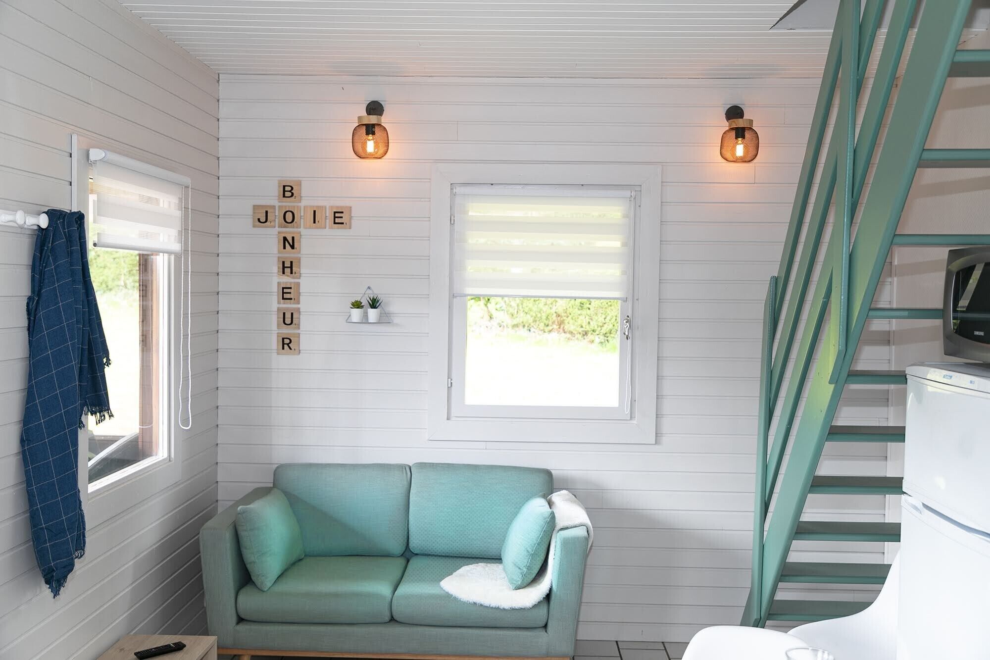 Ein gemütliches Zimmer mit einem türkisfarbenen Sofa, einem Fenster mit Jalousien, einer Treppe und einer Wand mit einer Leuchte sowie einem Schild mit dem Schriftzug 'BONJOUR'.
