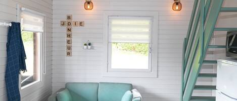 Een gezellige kamer met een blauwgroene bank, een raam met jaloezieën, een trap en een muur met een lamp en een bord waarop 'BONJOUR' staat.