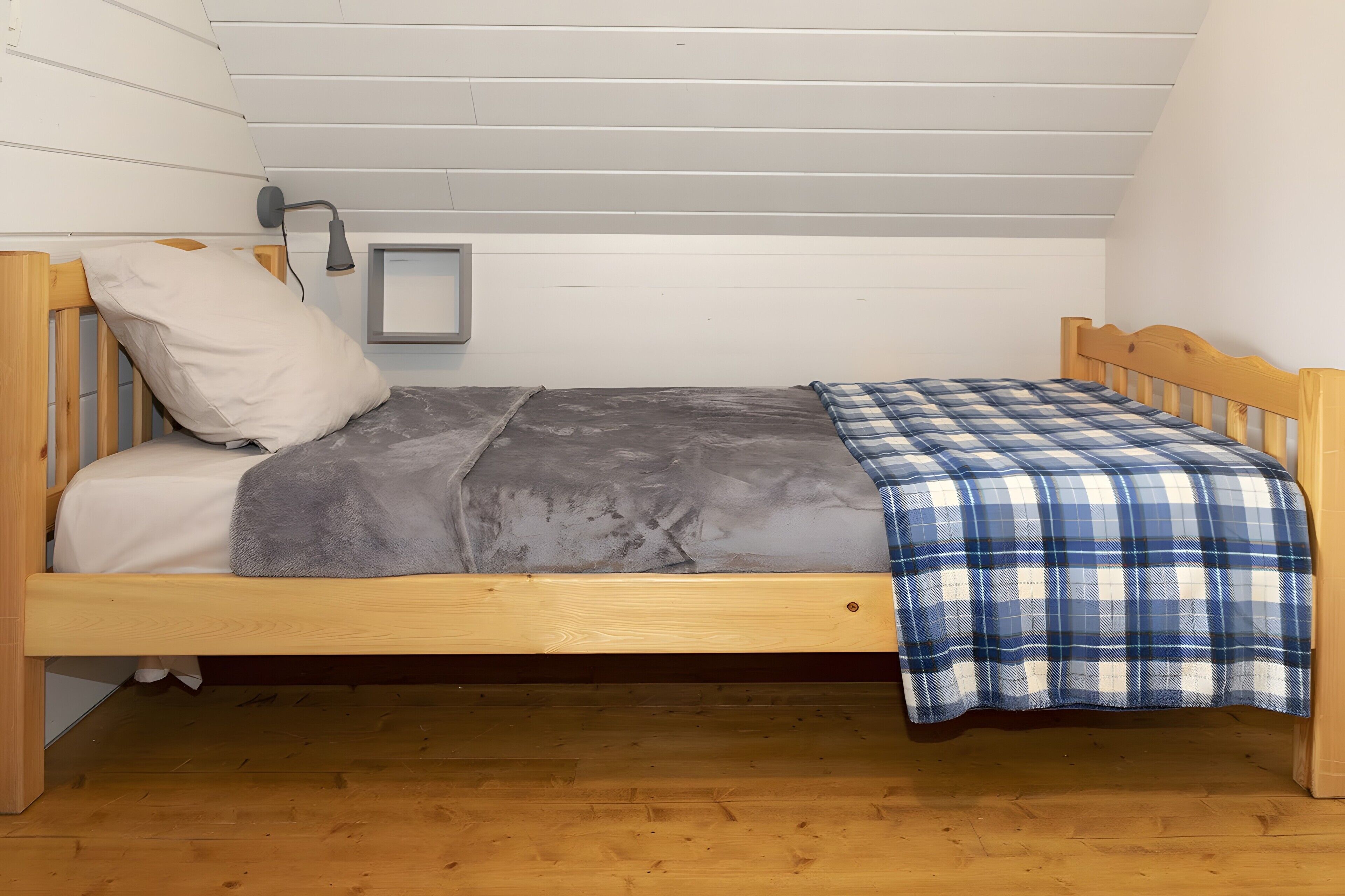 Un lit simple avec une couverture grise et une couverture à carreaux bleus et blancs, une tête de lit en bois et une lampe de chevet.