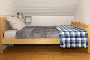 Un lit simple avec une couverture grise et une couverture à carreaux bleus et blancs, une tête de lit en bois et une lampe de chevet.