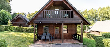 A wooden cabin with a balcony, a covered patio with seating, and a gravel driveway.