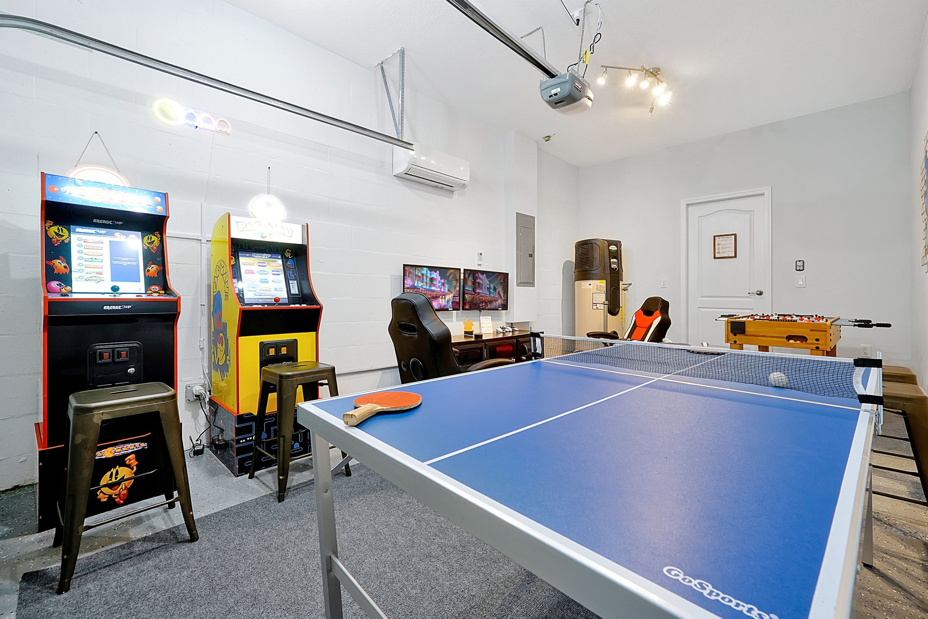 A room with a ping pong table, two arcade machines, a desk with a computer, and a foosball table.