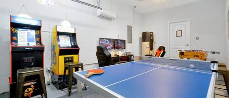A room with a ping pong table, two arcade machines, a desk with a computer, and a foosball table.