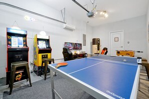 Ein Zimmer mit einer Tischtennisplatte, zwei Spielautomaten, einem Schreibtisch mit Computer und einem Kicker.