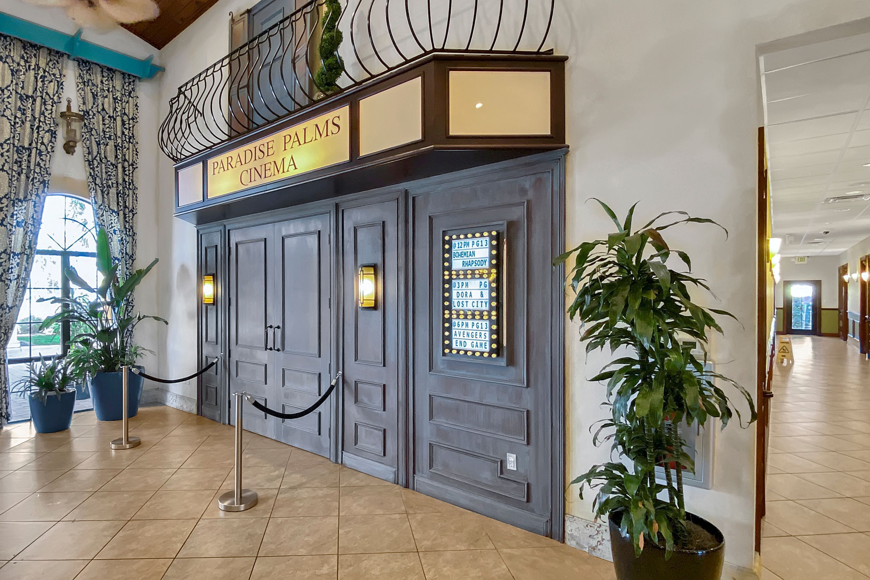Een bioscoop ingang met een bord waarop 'Paradise Palms Cinema' staat, een zwarte touwbalk en potplanten.