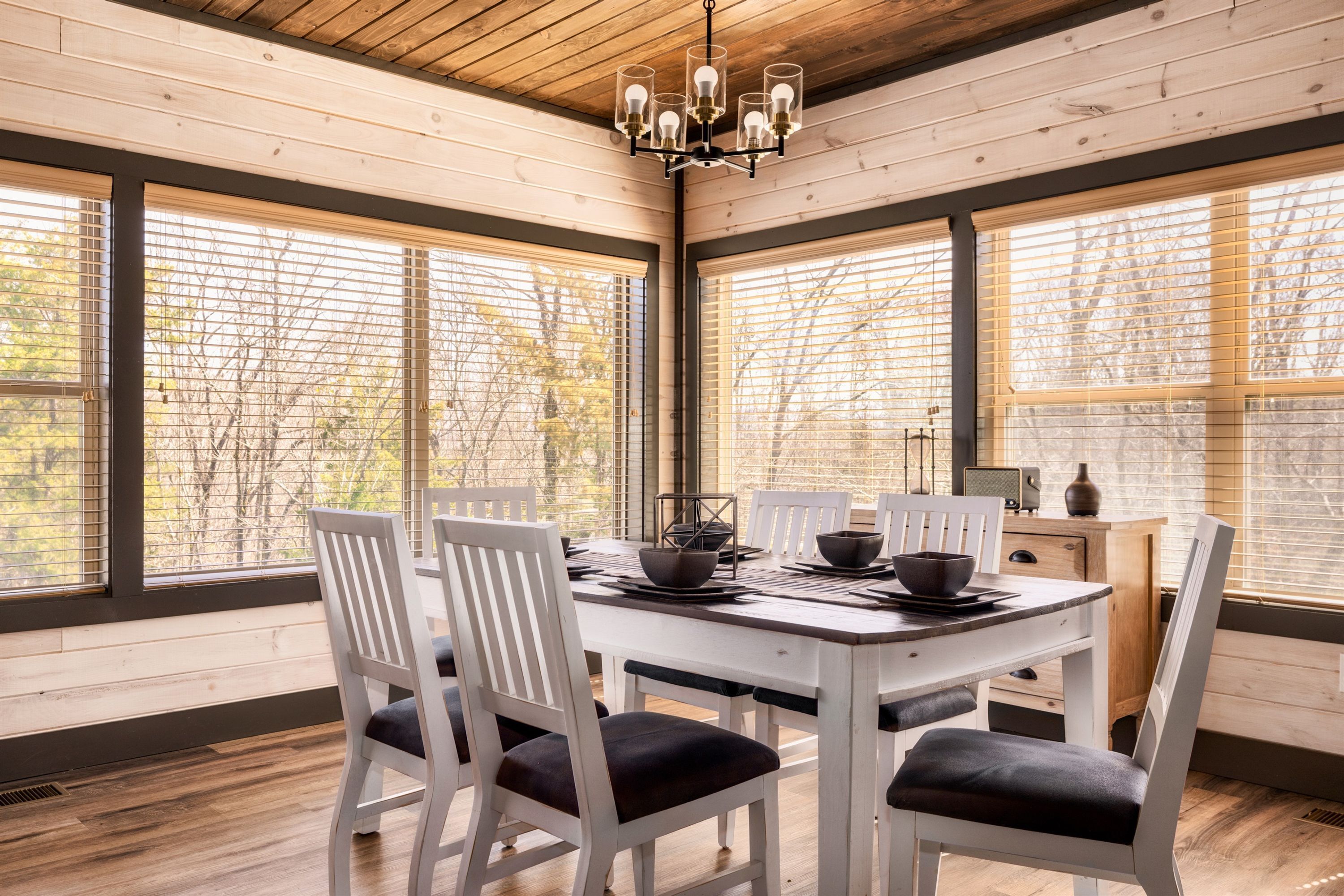 Un coin repas avec une table et des chaises en bois, de grandes fenêtres équipées de stores et un lustre.