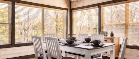 Un coin repas avec une table et des chaises en bois, de grandes fenêtres équipées de stores et un lustre.