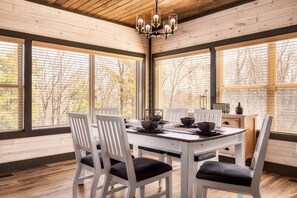 Un comedor con mesa y sillas de madera, ventanas grandes con persianas y una lámpara de araña.