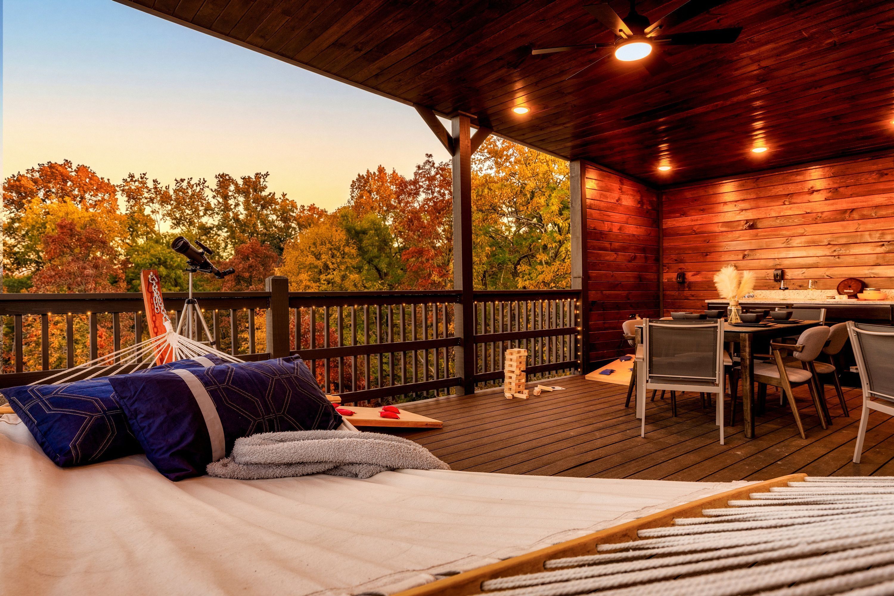 Eine Holzterrasse mit einer Hängematte, einem Tisch mit Stühlen und Blick auf Herbstbäume.