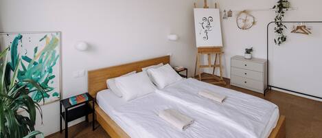A neatly made bed in a room with a wooden headboard, a small bedside table with a lamp, a wall-mounted easel with a painting, and a potted plant.