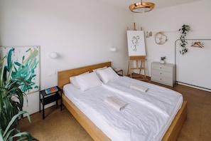 A neatly made bed in a room with a wooden headboard, a small bedside table with a lamp, a wall-mounted easel with a painting, and a potted plant.