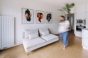 A modern living room with a white sofa, a kitchen area with a microwave and oven, and abstract art on the wall.