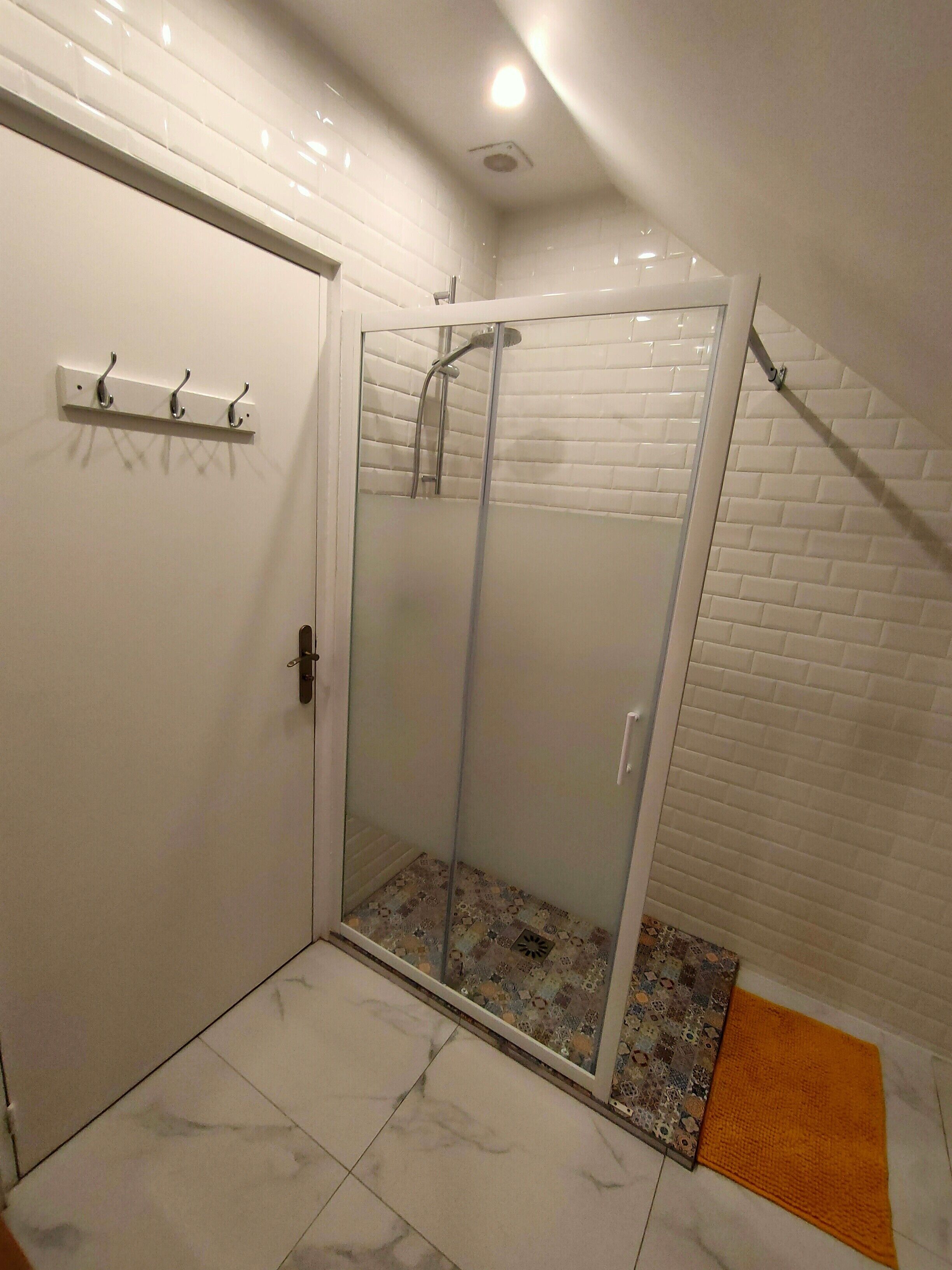 Une douche fermée par des parois de verre, avec un sol carrelé de blanc, une porte blanche dotée d’une poignée et un petit tapis orange.