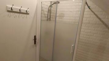 Une douche fermée par des parois de verre, avec un sol carrelé de blanc, une porte blanche dotée d’une poignée et un petit tapis orange.