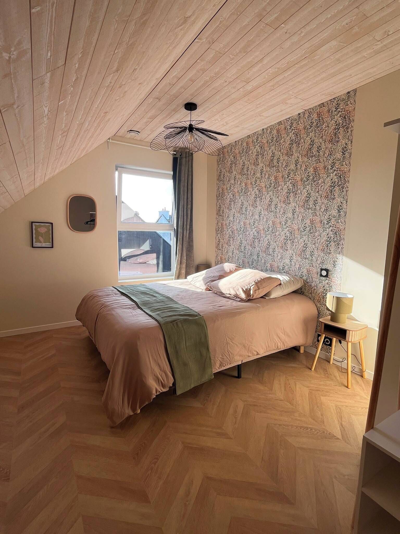 Ein Schlafzimmer mit Fischgrätparkett, einem Bett mit grüner Tagesdecke, einem gemusterten Wandpapier, einem Deckenventilator und einem Fenster mit Blick.
