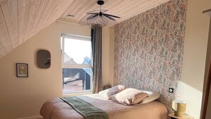 Une chambre avec un parquet en chevrons, un lit recouvert d’un plaid vert, un papier peint à motifs, un ventilateur de plafond et une fenêtre donnant sur un beau paysage.