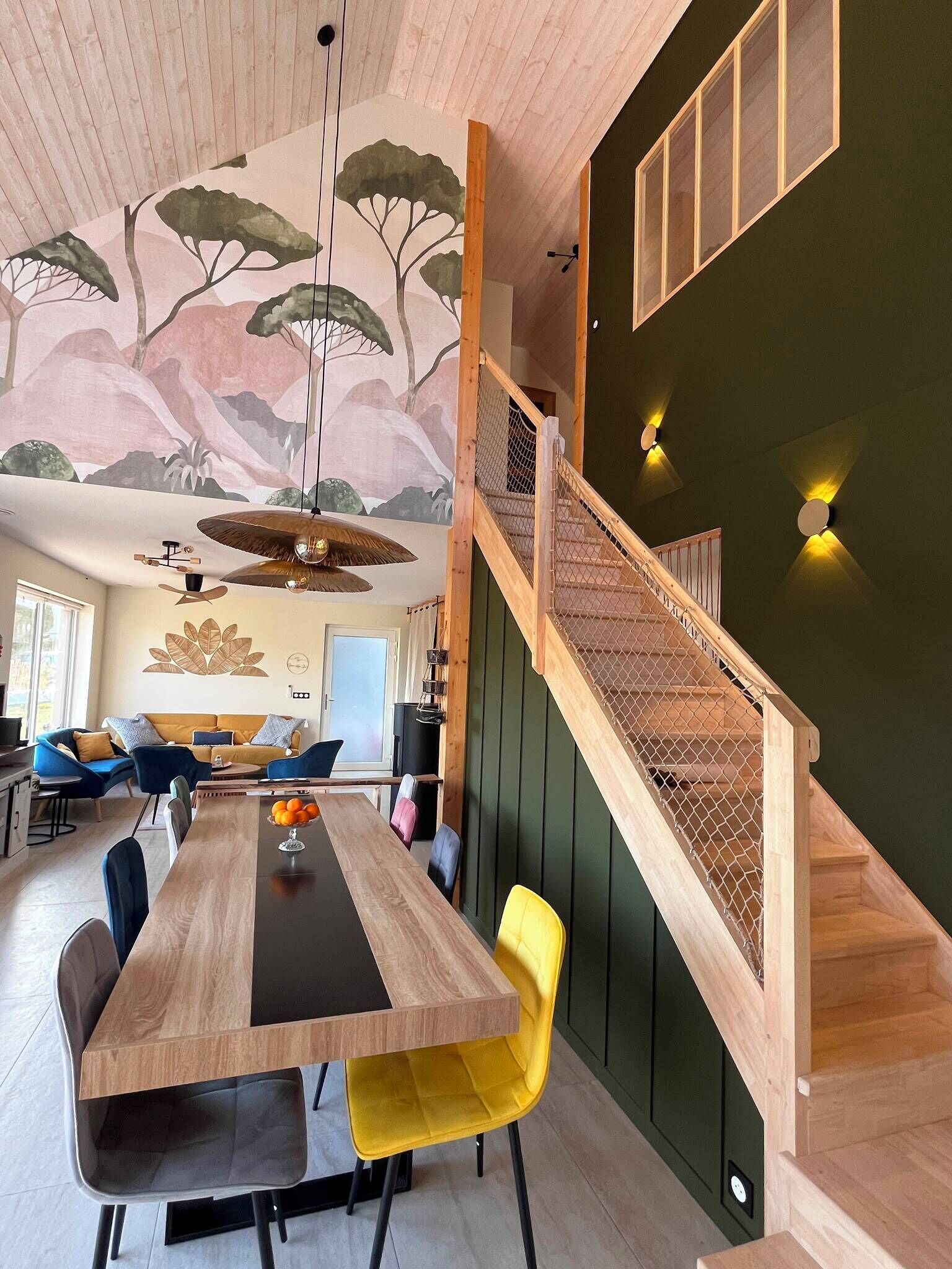 Un intérieur moderne avec une table à manger en bois, des chaises jaunes, un escalier avec une rampe en bois et une fresque murale.
