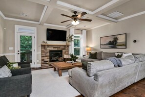 A living room with a fireplace, a ceiling fan, a flat-screen TV, and a large window. - Luxury Chapin Retreat | Pool, Theater, Gym + Bar (Chapin)