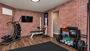 Un gimnasio en casa con una bicicleta estática, banca para pesas, pesas de caña y un televisor montado en la pared de ladrillo.