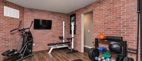A home gym with a stationary bike, bench press, kettlebells, and a TV mounted on the brick wall.