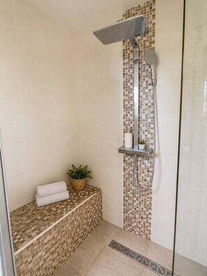 Eine Dusche mit Regendusche, Ablagefläche mit Pflegeartikeln und eine Sitzbank mit Pflanze und Handtüchern.