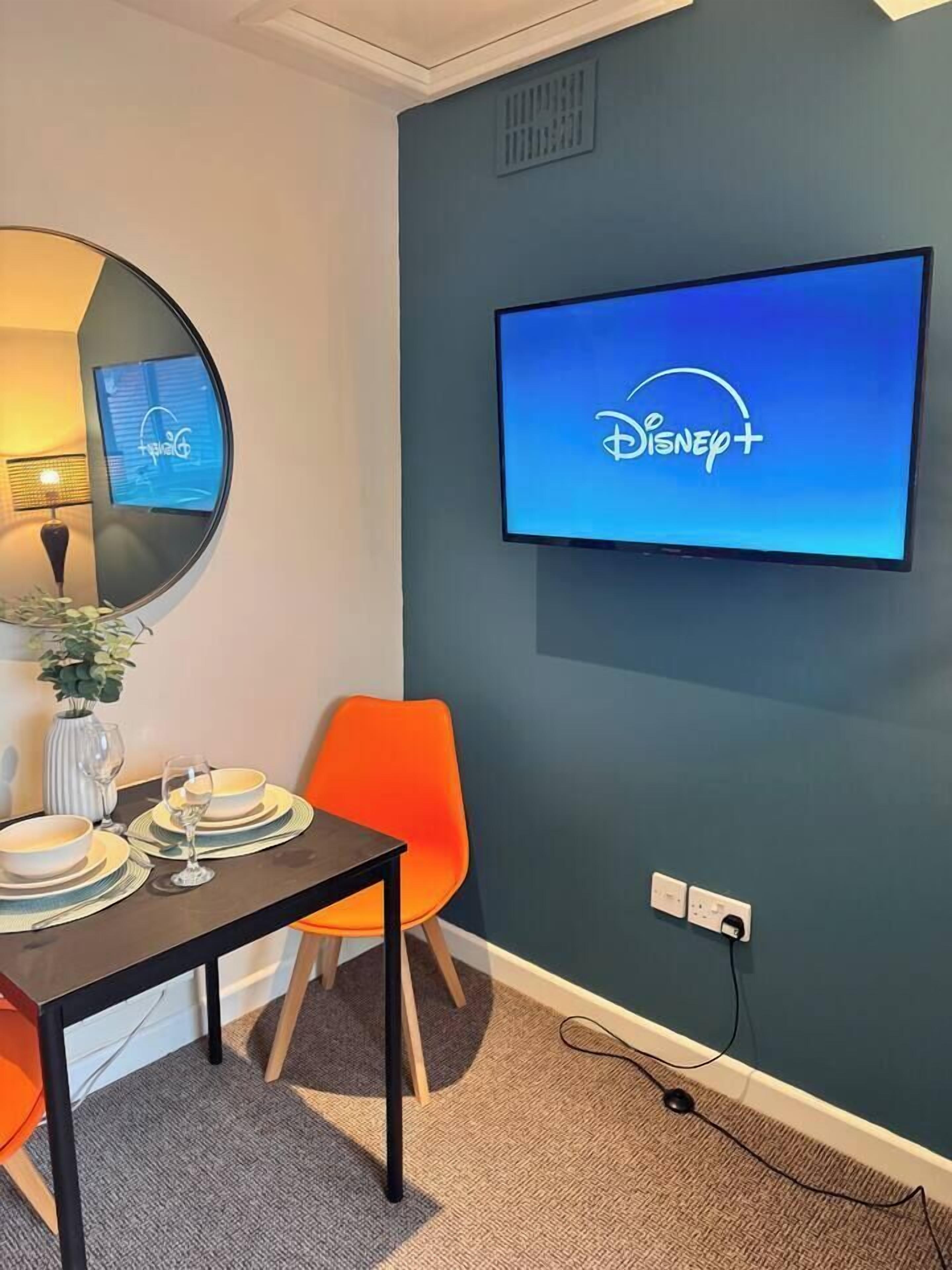 Une pièce avec un téléviseur affichant le logo de Disney+, une table à manger avec des assiettes et des verres, un vase avec une plante et une chaise orange.