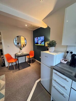 A compact kitchen with a refrigerator, a small dining table with two chairs, a coffee machine, and a flat-screen TV mounted on the wall. - #81 Stylish TopFloor|for 2|10 mins to Brueton Park (Shirley)