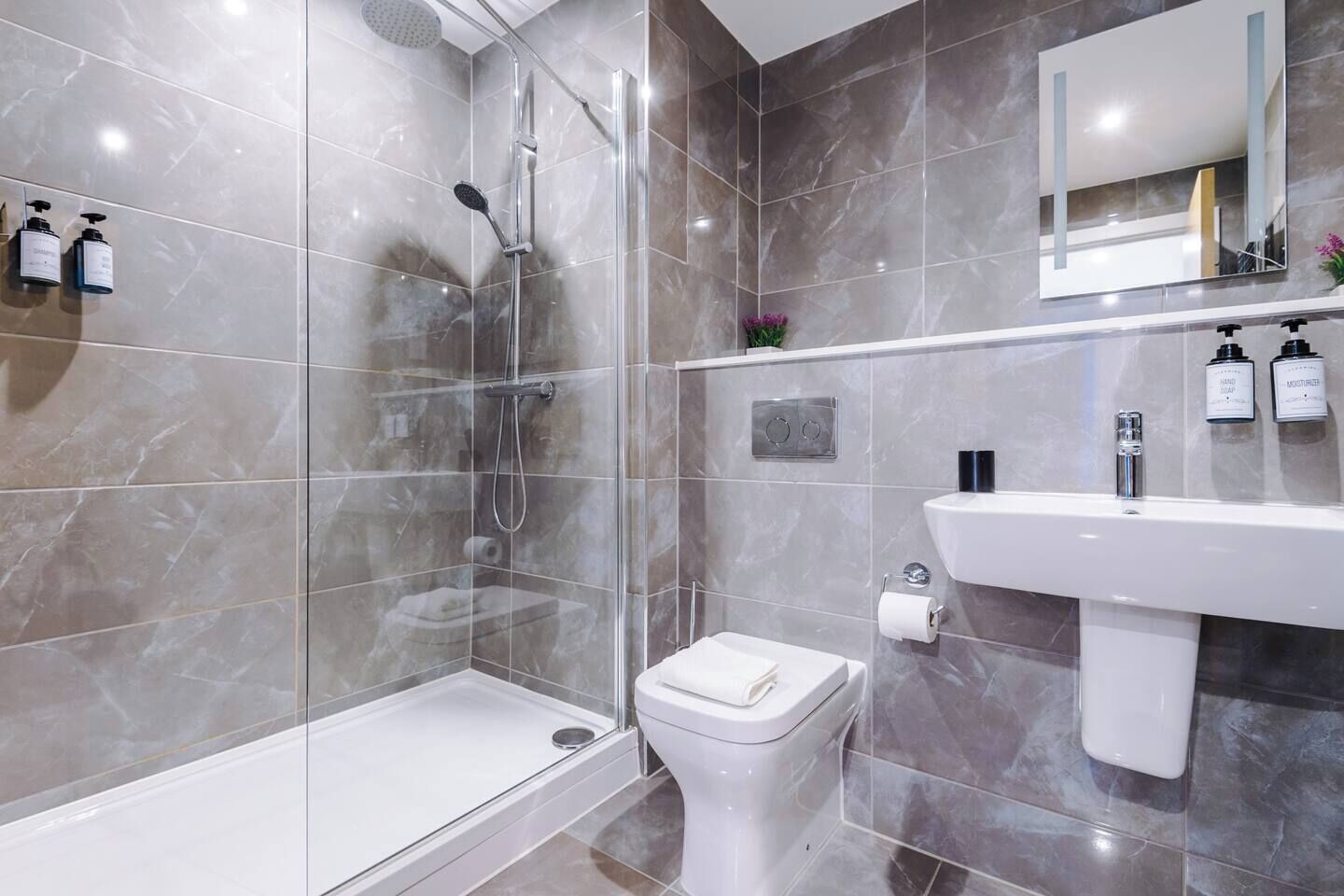Un baño moderno con ducha de vidrio, inodoro blanco y lavamanos con artículos de aseo.