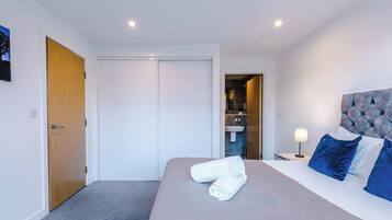 Quarto de hotel com cama, mesa de cabeceira, luminária, porta e vista para um banheiro.