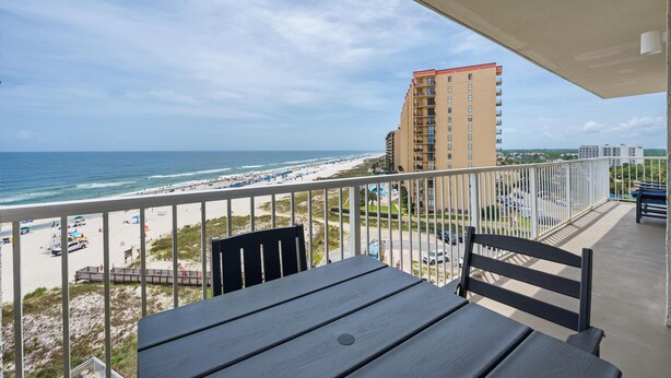 A balcony with a view of a beach and ocean, a tall building in the background, and a clear sky. - Seaside Beach & Racquet 3801 (Orange Beach)