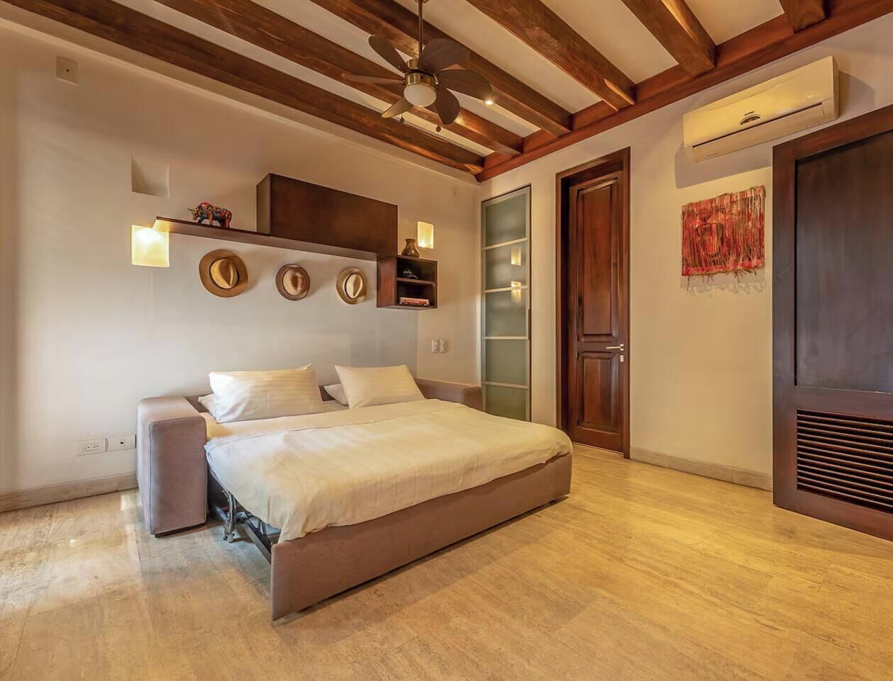 A bedroom with a bed, a nightstand, a wall-mounted shelf with decorative items, a door, and a wall-mounted air conditioning unit.