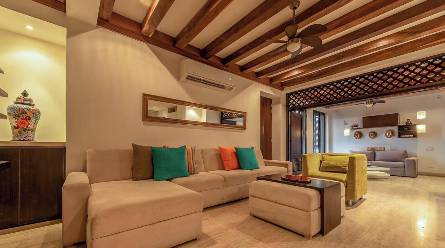 Ein Wohnzimmer mit Sofa, Hocker und Couchtisch. Eine Deckenleuchte, ein Spiegel und eine dekorative Vase stehen auf einem Schrank.