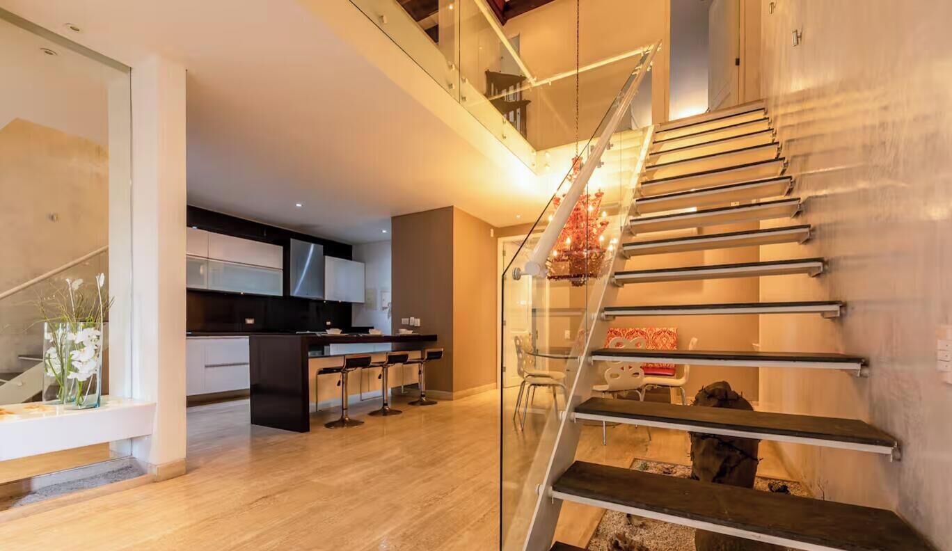 A modern kitchen with a central island and bar stools, a staircase with a metal railing, and a glass partition.