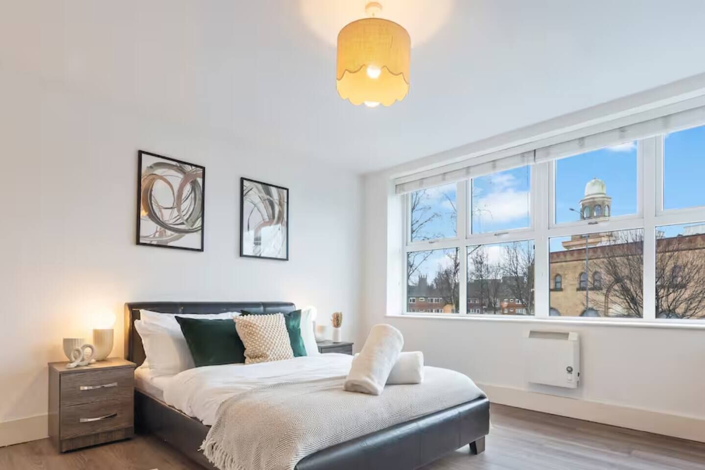 A bedroom with a large window, a bed, a bedside table, and two wall-mounted artworks.